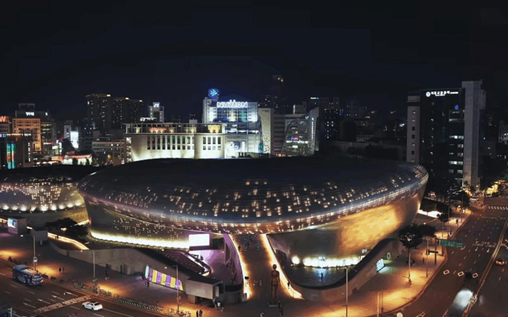ソウルの東大門デザインプラザ（DDP）の夜景を上空から撮影した写真。近未来的な曲線の建物がライトアップされ、周囲の街並みとともに鮮やかに輝いている。