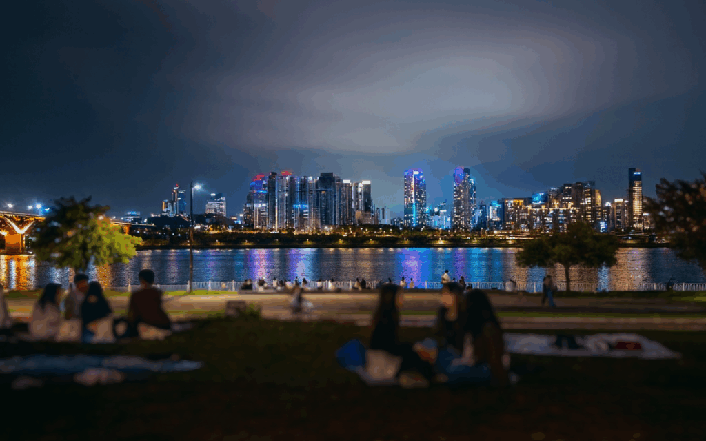 ソウルの漢江公園で川越しに高層ビル群の夜景を眺めながら、レジャーシートでくつろぐ人たちの様子。水面にはカラフルな光が映り込み、人気の夜デートスポットらしい落ち着いた雰囲気が広がっている。