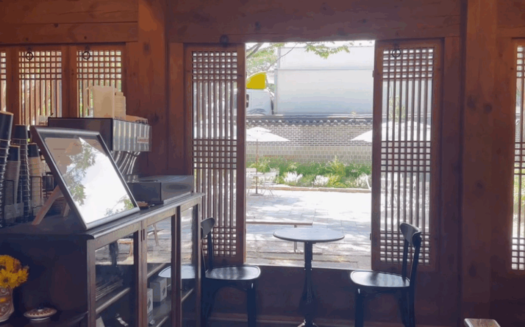 三清洞の韓屋カフェ店内から中庭を眺めたシーン。木の格子窓から光が差し込み、石畳のテラス席が見える落ち着いた雰囲気が魅力で、ソウル旅行で人気スポットとして訪れる人も多い。