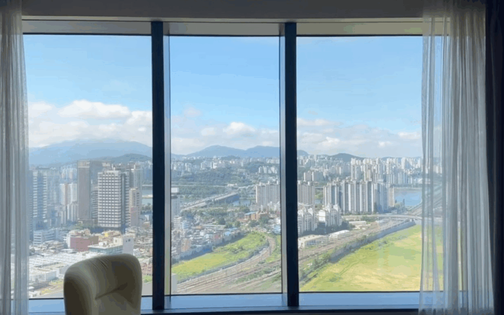 ソウル龍山区ノボテルスイーツアンバサダーホテル客室からの漢江（ハンガン）都市景観 晴天の青空と白い雲 高層ビル群と緑地帯 遠景に山並み HYBEビル・龍山駅周辺エリア K-POP推し活旅行の5つ星スイートからの眺望