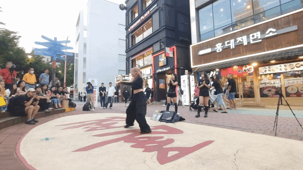 ソウル弘大で行われているK-POPダンスのストリートパフォーマンスを、若者や観光客が囲んで楽しむ夕方の風景。